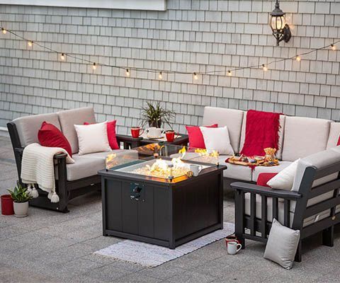 Outdoor patio with seating around a lit fire pit, string lights, red and white pillows, and a grey house.