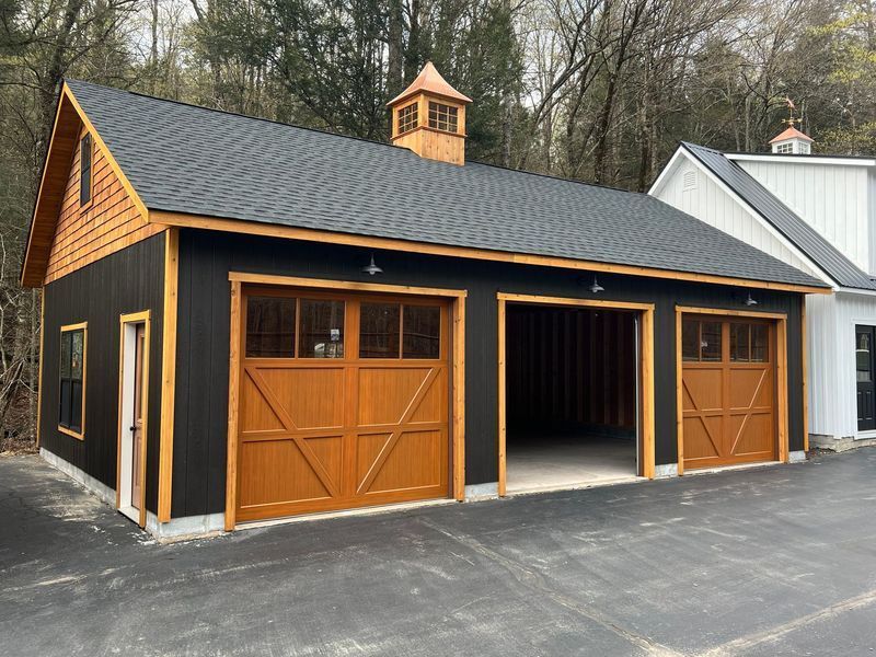 Black and brown three-bay garage with wooden doors. Small cupola atop. Set in a natural area.
