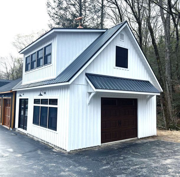 White two-story barn-style building with black roof and trim. Features a dark brown garage door and several windows.