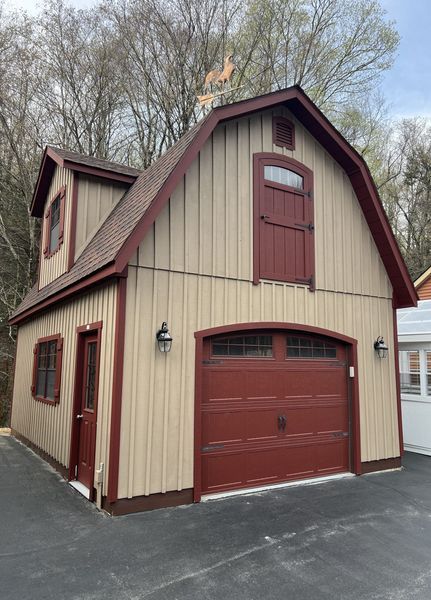 Tan and maroon two-story barn-style garage with a dormer, windows, and a closed garage door, on a paved surface.