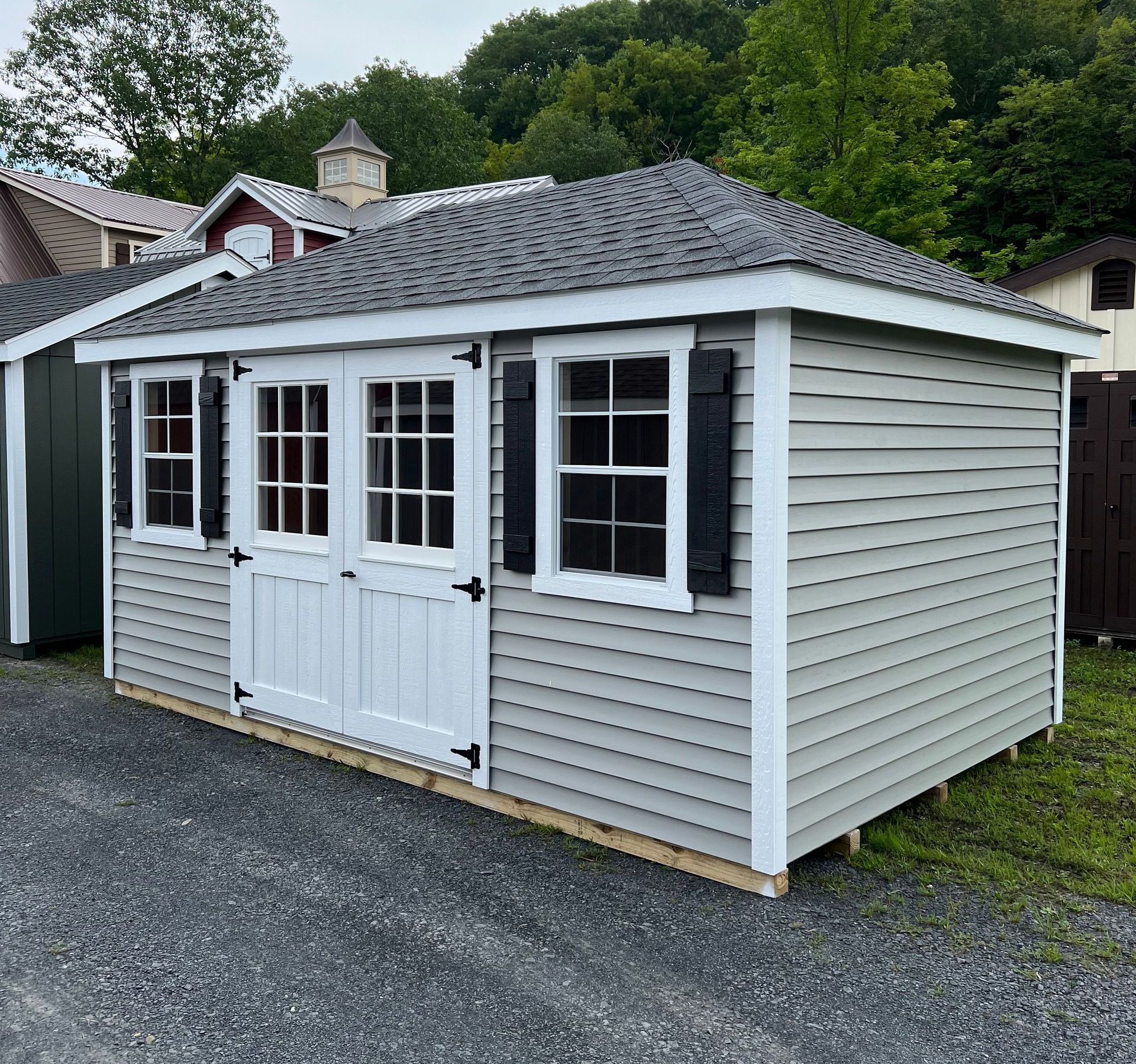 Gray-sided shed with white doors, windows, and trim; black shutters, gray roof, gravel ground.