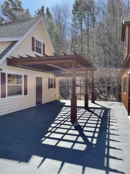 Yellow house with a brown pergola casting a shadow on a black asphalt patio. Sunny day.
