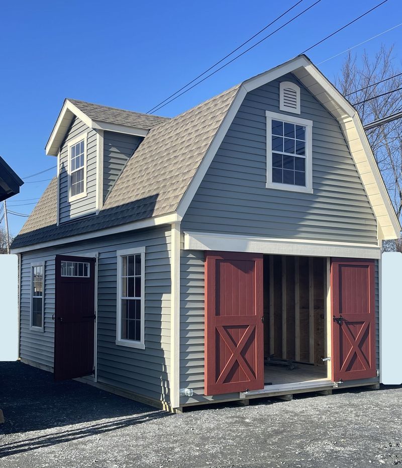 Two-story gray shed with red doors, tan roof, and blue sky.