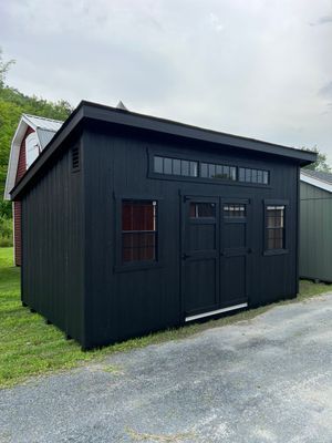 Black shed with a slanted roof, windows, and double doors, sitting on grass with a gravel path.