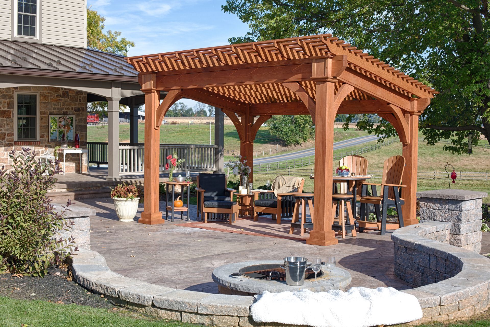 Brown wooden pergola over patio with seating, fire pit, and stone wall, next to a house.
