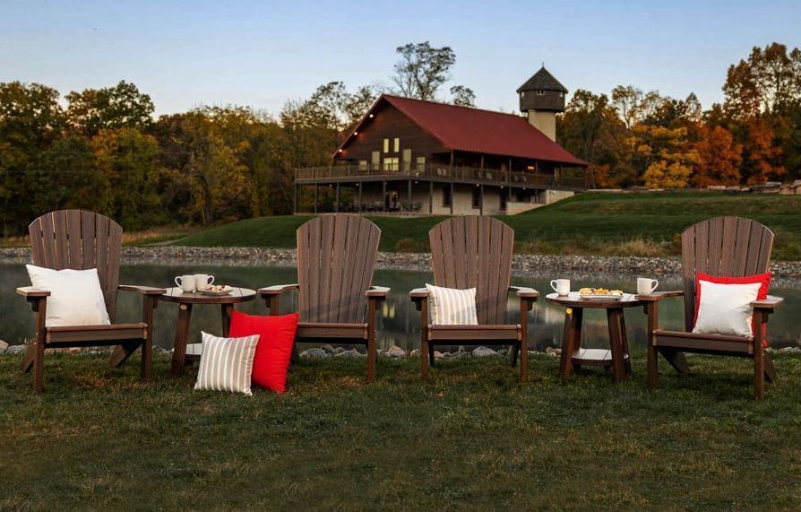 Four wooden Adirondack chairs with pillows and small tables on a grassy bank overlooking a pond and a house.