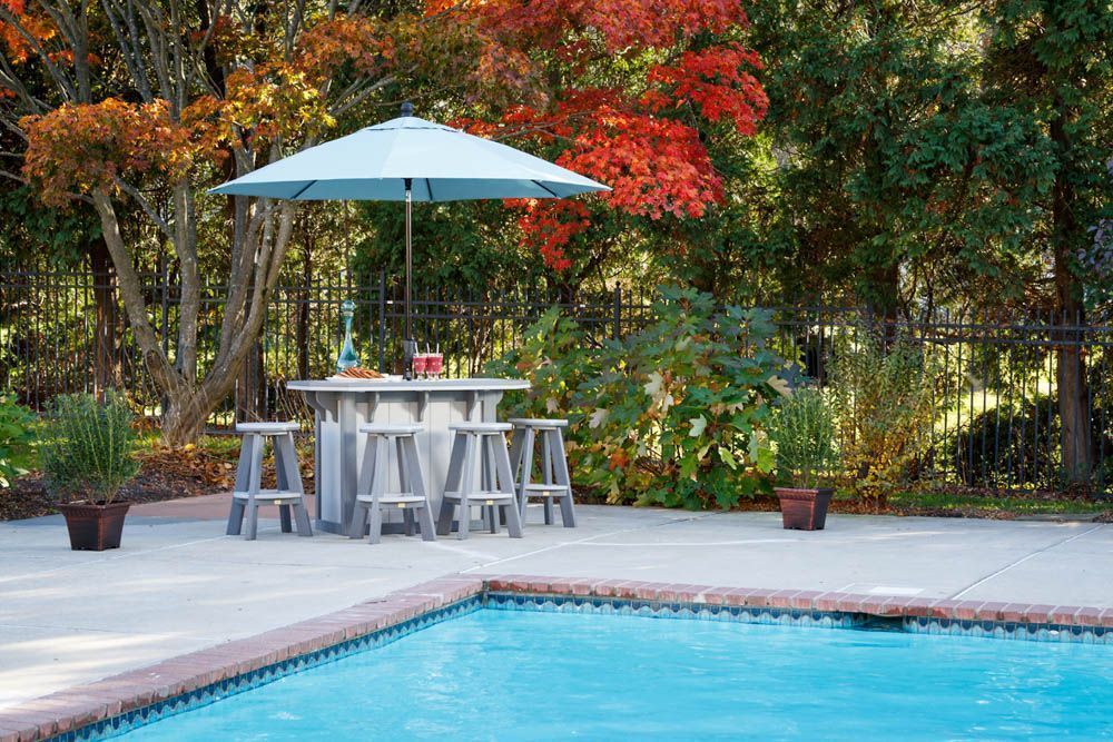 Backyard pool scene with a bar table, stools, and umbrella under trees with fall foliage.