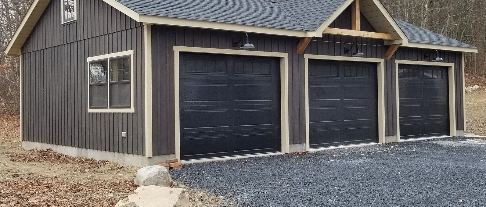 A three-bay garage with black doors, gravel driveway, and a wooded backdrop.