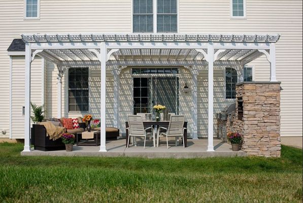 White pergola over patio with outdoor furniture, next to a stone column, house in background.