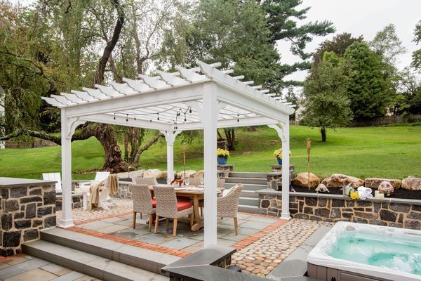 White pergola over outdoor dining set on stone patio, with a hot tub and a grassy yard in the background.
