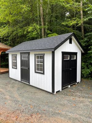 White shed with black trim and doors; two windows, gravel ground, trees in background.