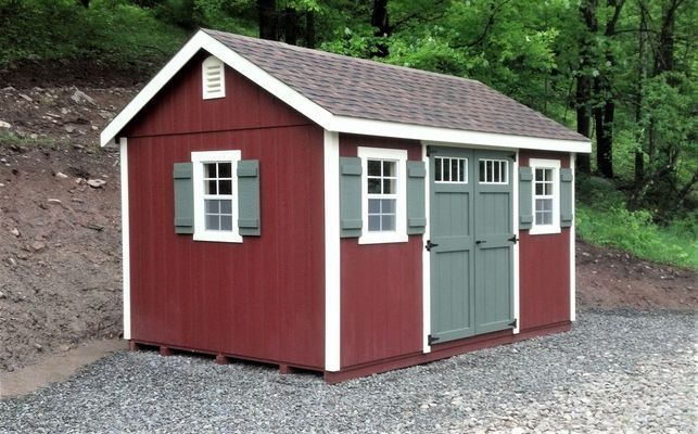 Red storage shed with white trim and green doors, gravel base, set against a wooded background.