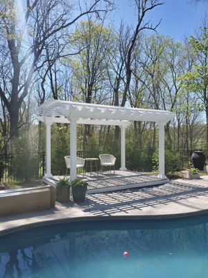 White pergola with seating by a pool; sunny day, trees in background.