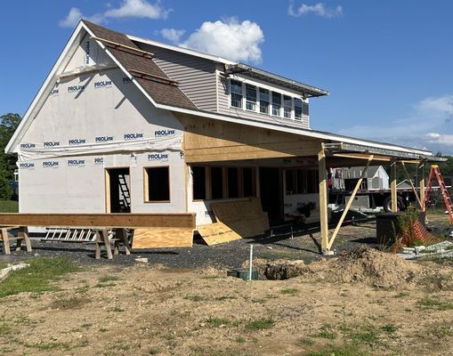 Construction site: two-story house with attached porch, siding and roofing in progress. Blue sky, sunny day.