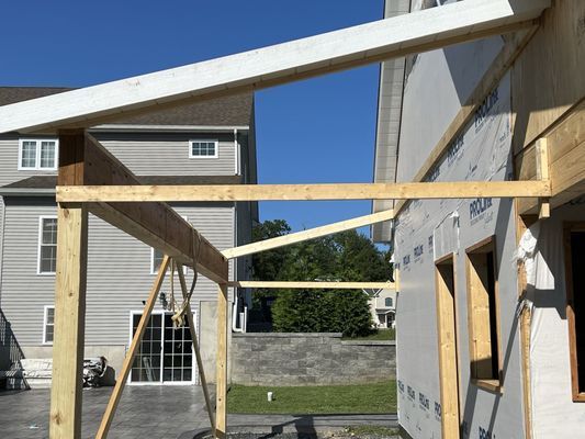 Wooden frame of an outdoor structure under construction next to a building; blue sky.