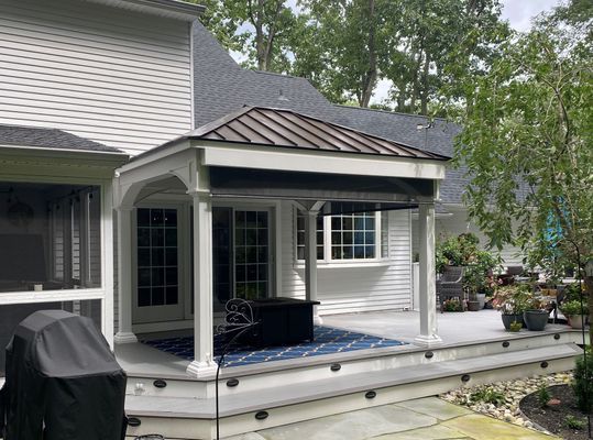 Backyard deck with a gazebo and outdoor seating. Gray deck, white house, green trees.