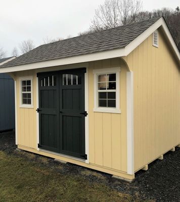 Yellow shed with dark green double doors and white-framed windows; set outdoors on gravel.