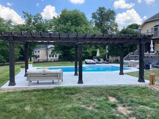 Pergola over concrete patio beside a pool with seating, trees, and a house in the background.