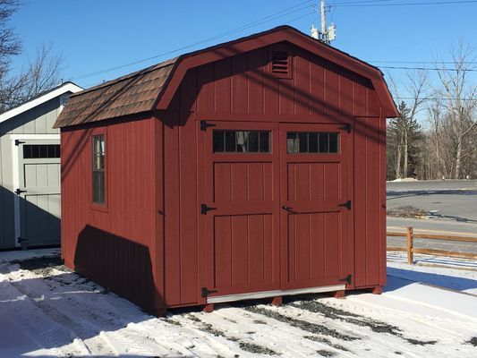 Red wooden shed with a barn roof and double doors, sitting on snow, against a blue sky.