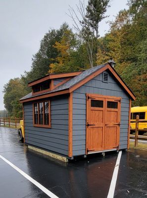 Small gray shed with brown trim and doors in a parking lot on a rainy day.