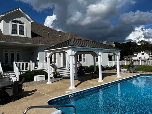 White gazebo by a pool next to a large white house with dark roof. Blue pool water. Cloudy sky.