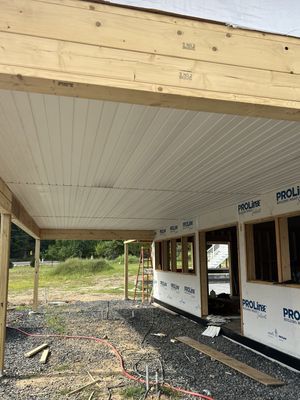 Construction of a building's porch with white panel ceiling and exposed wooden beams. Proline wrap on walls, gravel ground.