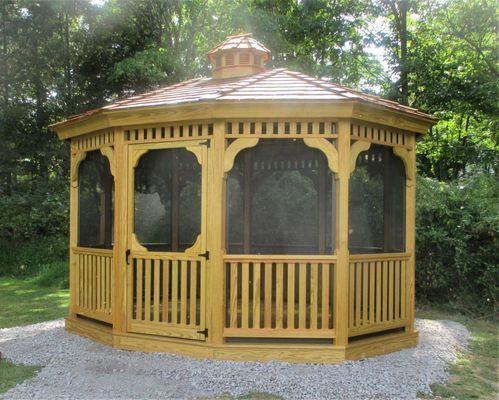 Wooden gazebo with brown roof, screened windows, on a gravel base, surrounded by greenery.