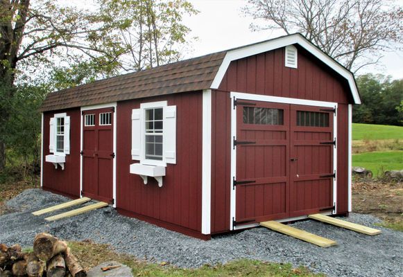 Red shed with white trim and windows, two doors, brown roof, gravel base, in a grassy field.
