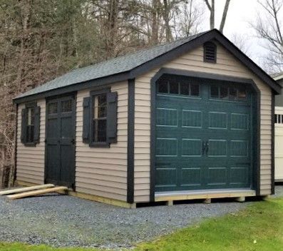 Tan shed with dark green garage door, shutters, and trim, gravel ground.