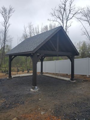 Dark brown wooden carport with a gray roof, standing on concrete pads, gravel ground, white fence in the background.