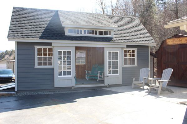 Small blue shed with white-framed windows and doors, Adirondack chairs on a patio, and a car in the background.