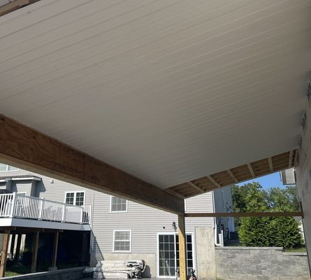 A view from under a porch roof with a light-colored ceiling, beams, and a grey house visible in the background.