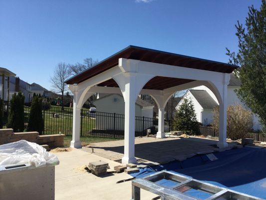 White pergola with arched supports over a pool; blue cover; brown roof. Sunny, residential setting.