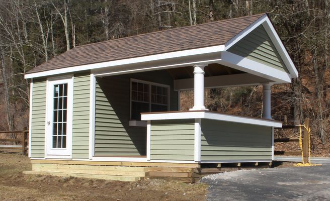 Small green building with covered porch; brown roof, white trim. Situated near a road and trees.