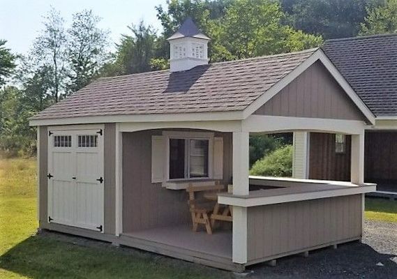 Tan shed with white doors and trim, attached porch, picnic table, and cupola.