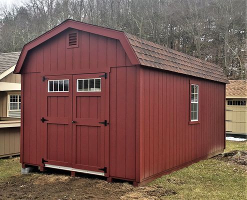 Red barn-style shed with double doors, small window, and brown shingle roof on grass.