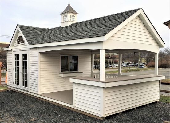 White outdoor shed with a bar-like counter, porch, and cupola. Set on gravel.
