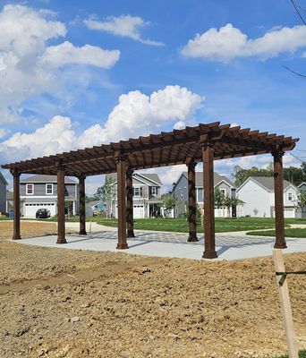 A brown pergola over a concrete pad with houses in the background, under a cloudy blue sky.