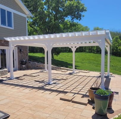 White pergola over a brick patio. Green grass and house in background. Sunny day.