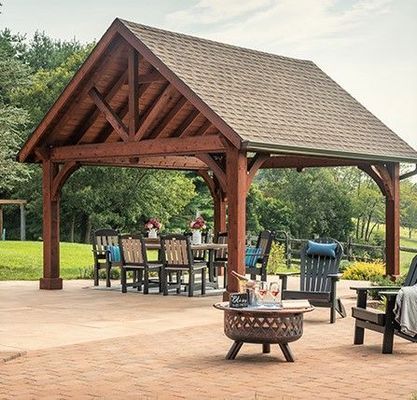 Wooden gazebo with dining set and chairs on a patio, fire pit in foreground, trees in background.