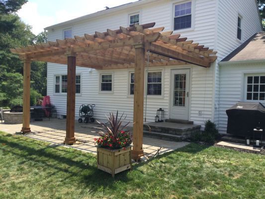 Wooden pergola attached to a white house, over a patio with a flower box and grill.
