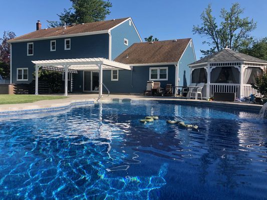 Backyard with blue pool, blue house, white pergola, and gazebo. Sunny day.