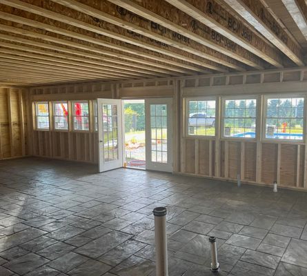 Interior of a room under construction with exposed wooden beams, windows, and a partially finished floor.