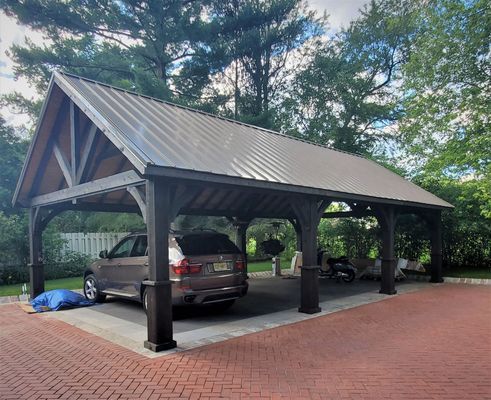 Car parked under a dark wood carport with a metal roof, in a brick driveway.