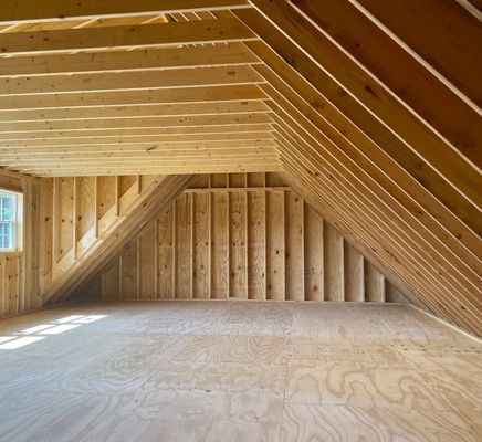 Interior of an unfinished attic space, with wood framing and plywood flooring, illuminated by sunlight.