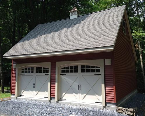 Red two-car garage with cream-colored doors and gray shingle roof in a wooded area.