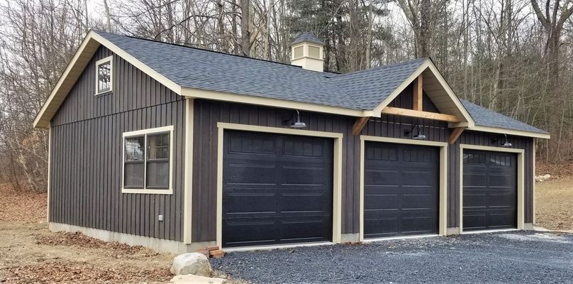 Dark-brown garage with three bays and a matching roof, set in a wooded area.