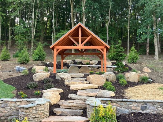Wooden gazebo with stone steps, surrounded by landscaping and trees.