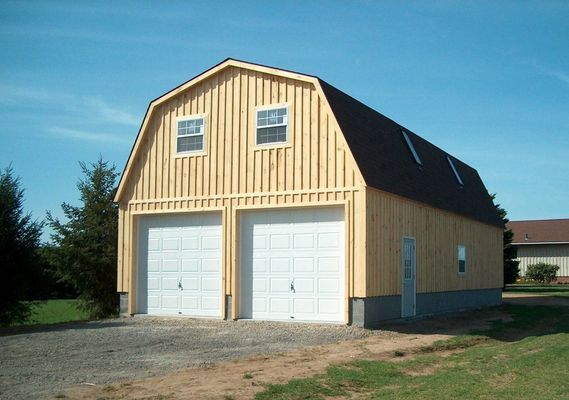 Two-story wooden barn with two garage doors, windows, and a dark roof on a sunny day.