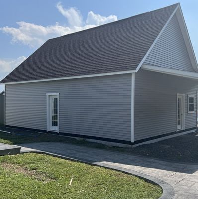 Gray building with a dark roof, white trim, and two doors next to a grassy yard and patterned sidewalk.
